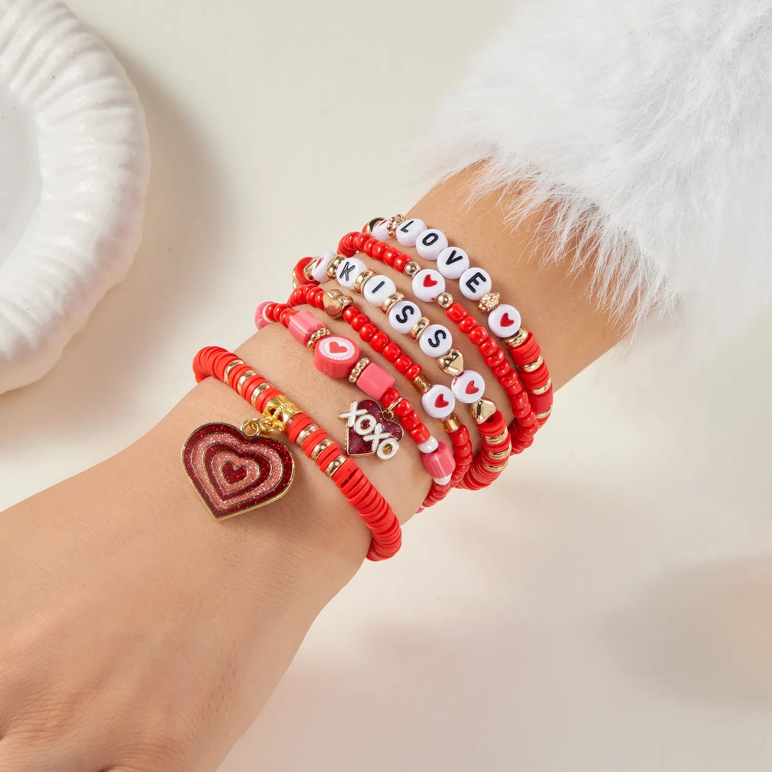 Hand wearing multiple beaded bracelets with heart and letter charms on a light background
