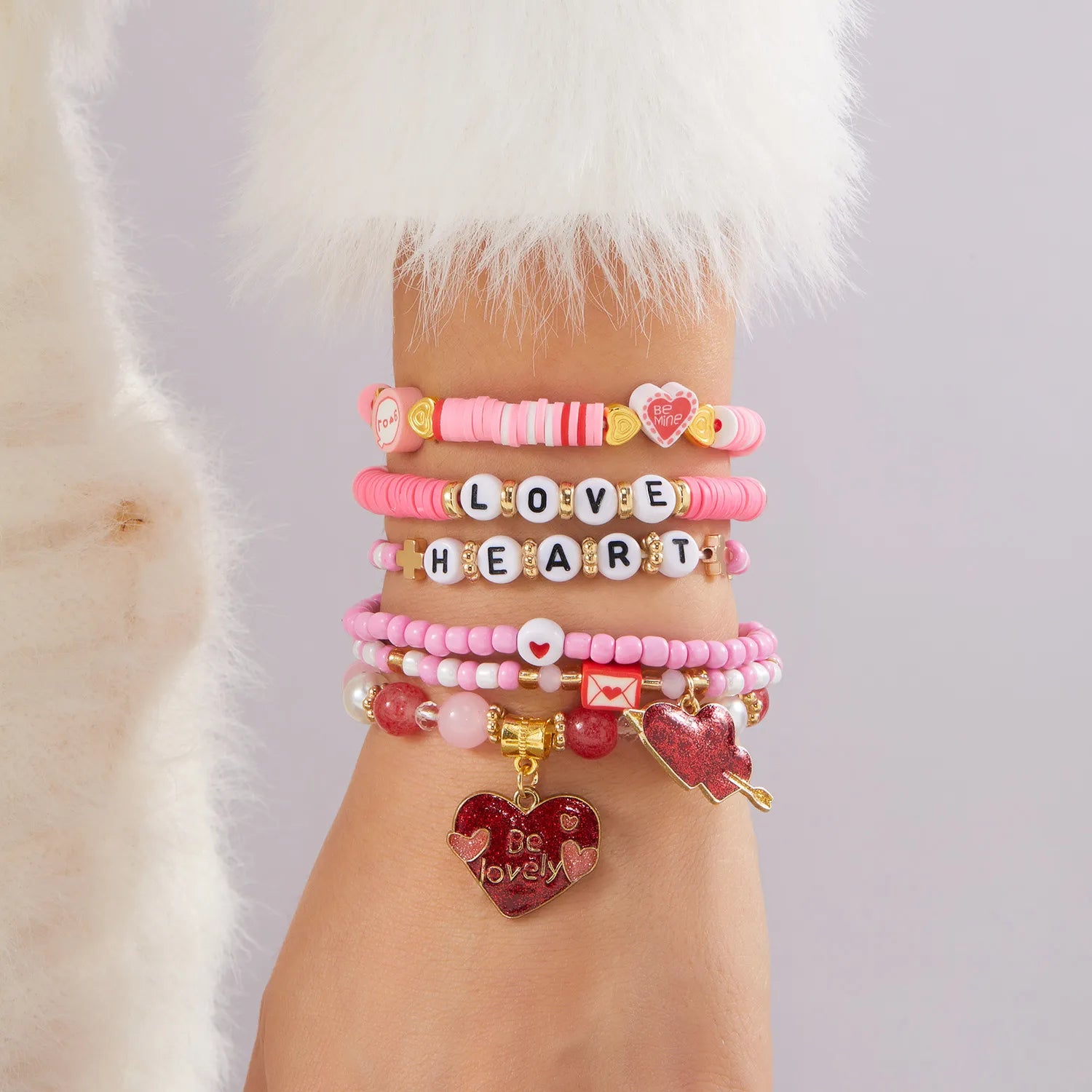 Multiple pink bracelets with heart and letter charms on a wrist, wearing a white fur coat.