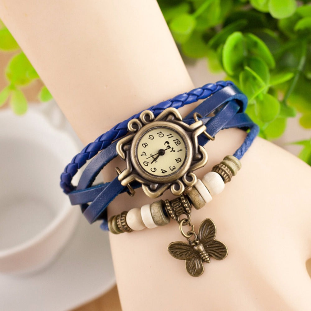 Blue braided bracelet with a vintage-style watch face and butterfly charm on a wrist against a blurred green background.