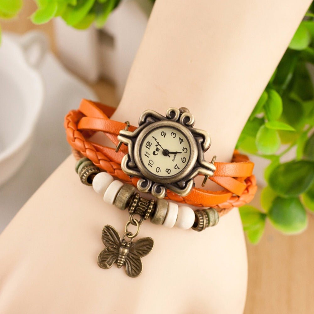 Wrist wearing a bracelet with a watch face and butterfly charm, against a blurred natural background.