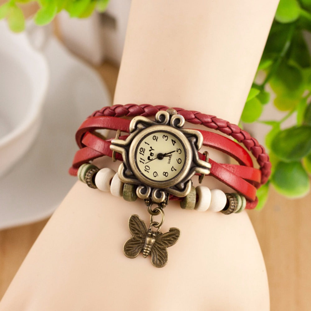 Red braided bracelet with a vintage-style watch and butterfly charm on a wrist, blurred background