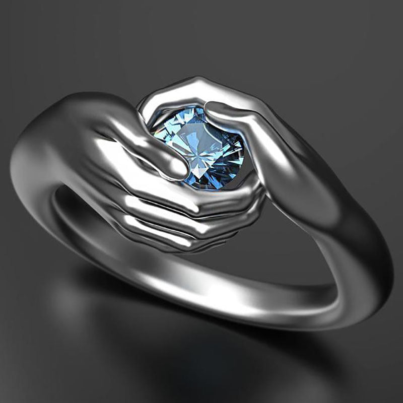Silver ring with a blue gemstone held between two hands on a dark background