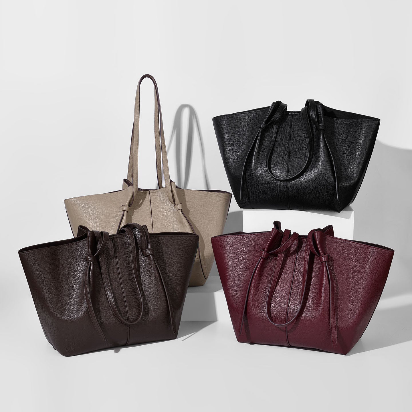 Four leather tote bags with drawstring handles in black, Khaki, chocolate, and burgundy, arranged on white pedestals against a light gray background.