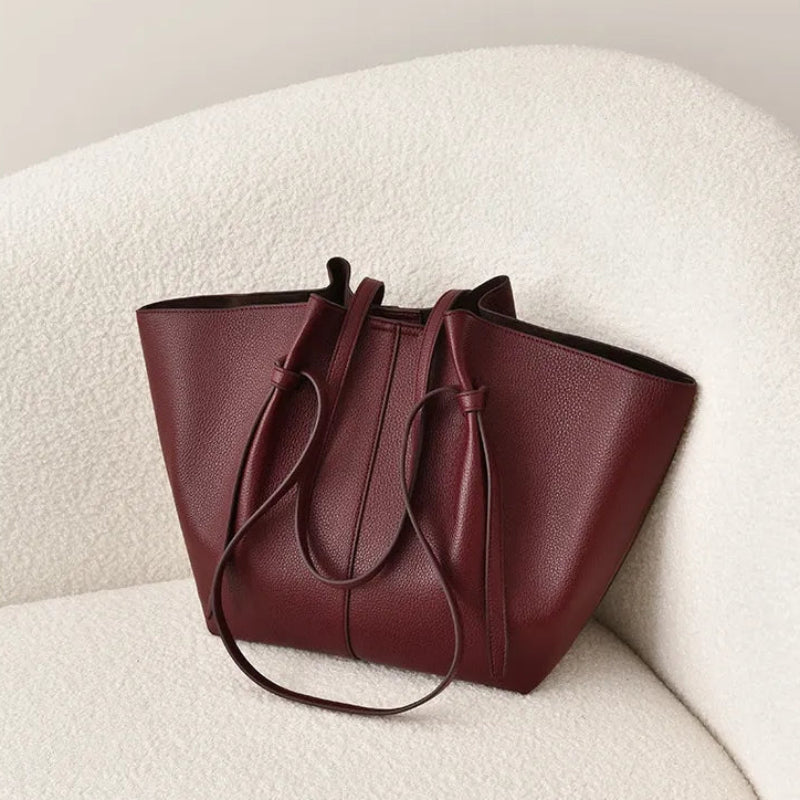 A burgundy pebbled leather tote bag with a long shoulder strap, resting on a textured cream boucle chair against a neutral beige background.