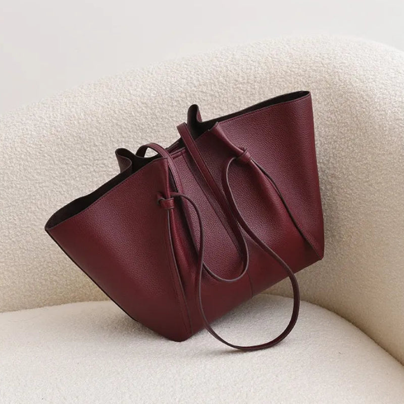 A structured tote bag in deep burgundy faux leather, styled on a cream-colored textured chair against a neutral light gray background.