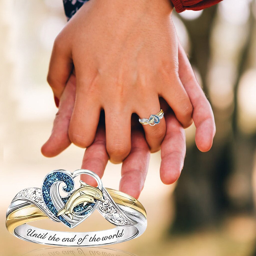 Two hands holding together with a heart-shaped ring featuring blue and gold colors and an engraved message.